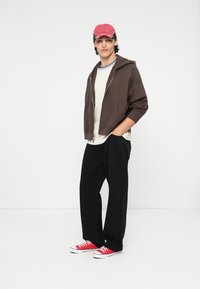 Brun zip-up hettegenser, hvit t-skjorte, svarte vide bukser og røde sneakers. Kappen er rosa med et logo. Modellen står foran en ensfarget bakgrunn.