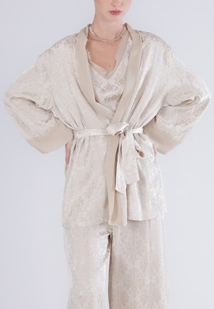 Žena v béžovém hedvábném vzorovaném županu se širokými rukávy a odpovídajícími kalhotami, zavázanými v pase páskem, s rukama v bok.