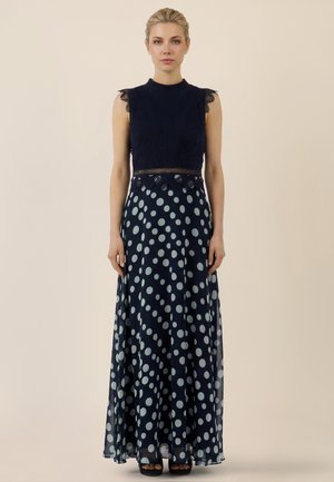 Woman standing facing forward wearing a sleeveless navy dress with lace top and large white polka dots on a flowing skirt.