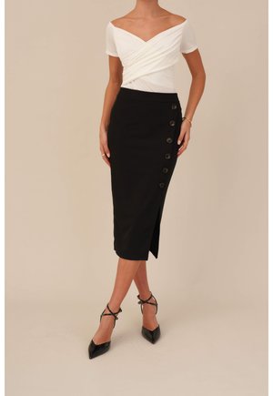 Falda lápiz negra con detalles de botones laterales, combinada con una blusa blanca de hombros descubiertos. Los zapatos son de charol negro con tiras en el tobillo.