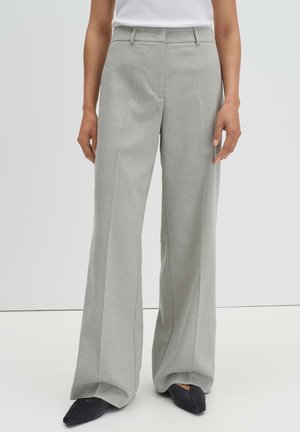 Femme portant un pantalon large gris clair et des chaussures noires à bouts pointus, debout devant un fond clair uni.
