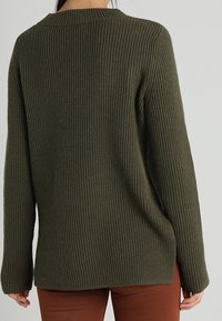 Person, der einen olivgrünen gerippten Pullover und braune Hosen trägt, von hinten vor einem schlichten, hellen Hintergrund gezeigt.