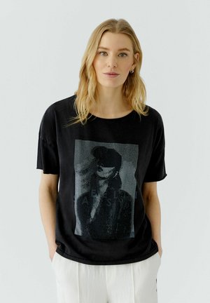 Blonde Frau trägt ein schwarzes Grafik-T-Shirt mit monochromem Porträt-Print und weißen Hosen, steht mit den Händen in den Taschen vor weißem Hintergrund.