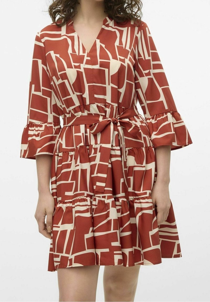 Femme portant une robe rouge et beige à motifs géométriques mi-longue, avec des manches trois-quarts à volants et une ceinture nouée à la taille.