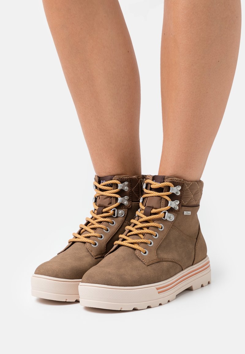 Bottines en daim marron avec col matelassé, lacets jaunes, œillets argentés, et une semelle en caoutchouc clair ornée de rayures horizontales colorées.