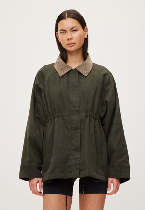 Femme portant une veste oversize vert foncé avec col marron et taille à cordon, associée à un short noir, debout devant un fond uni.