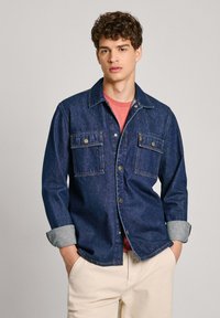 Camicia di jeans blu scuro con due tasche sul petto, chiusura a bottoni e polsini in azzurro contrastante, indossata sopra una maglietta rossa.