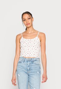Top cropped branco com padrão floral vermelho, alças finas estilo spaghetti e bordas recortadas, combinado com jeans de cintura alta em azul claro.