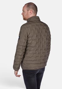 Giubbotto imbottito in verde oliva, con un motivo trapuntato, colletto alto e chiusura con zip. Accento nero sulla spalla. Indossato con jeans scuri.
