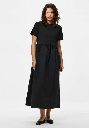 Frau mit lockigem Haar, die ein schwarzes Midi-Kleid mit kurzen Ärmeln und schwarze Ballerinas trägt, steht vor einem schlichten weißen Hintergrund.