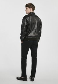 Czarna skórzana kurtka bomberka z prążkowanym dołem i mankietami, noszona na czarnych spodniach i skórzanych butach, pokazana z tyłu. Gładka tekstura.
