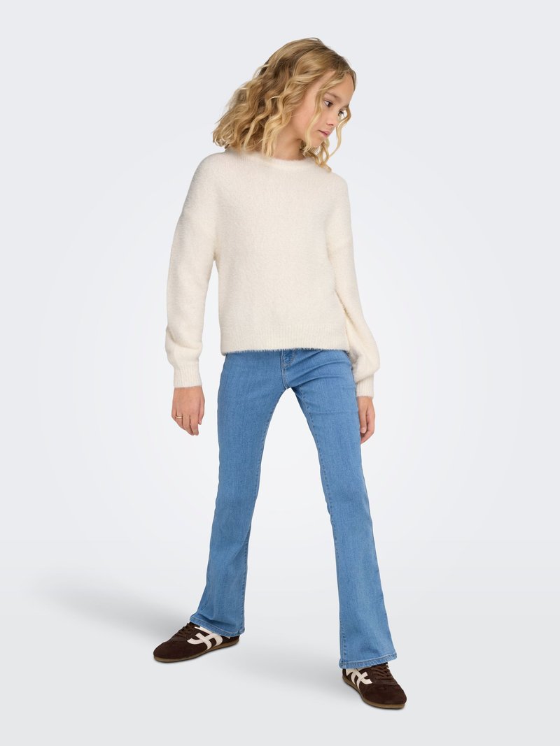 Cremefarbener, gerippter Strickpullover mit langen Ärmeln, kombiniert mit hellblauen Schlagjeans und braunen Sportschuhen mit weißen Akzenten.