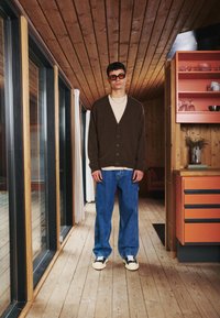 Brown cardigan over beige top, paired with baggy blue jeans and white sneakers. Model wears brown sunglasses, stands in a wooden interior.