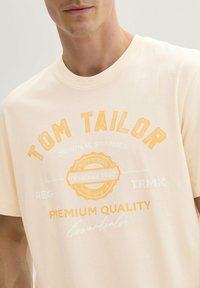 Kerge virsiku värvi T-särk, millel on kaubamärk "TOM TAILOR" julge oranži tekstiga. Särk sisaldab ümmargust logodetaili, millel on kiri "alates 1962" ja "premiaalne kvaliteet."
