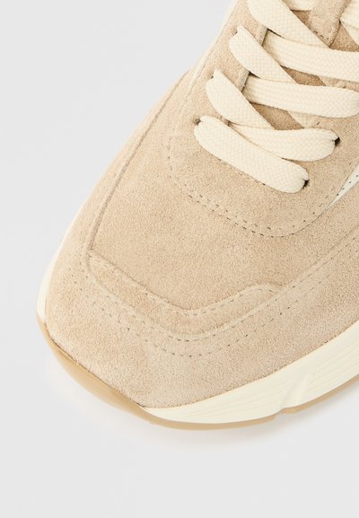 Μπεζ σουεντ αθλητικό παπούτσι με μπεζ κορδόνια, διαθέτει υφή επιφάνειας και στρογγυλεμένη μύτη. Σημαντικές ραφές κατά μήκος της πλευράς.