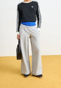 Osoba oblečená v černém dlouhém tričku Adidas, s vrstveným modrým páskem, šedými širokými kalhotami, držící černou tašku, stojící na žluté podlaze.