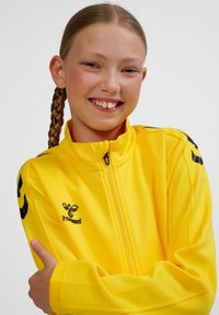 Gul zip-up jacka i slätt tyg, med svart logotyp och detaljer, hög krage och två ränder på varje ärm.