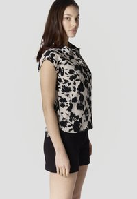 Mladá žena s dlhými hnedými vlasmi, oblečená v čierno-bielej kvetinovej bezrukávovej blúzke a čiernych šortkách, stojaca bokom na jednoliatom pozadí.