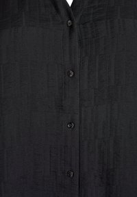 Melna pogājamā krekla ar teksturētu audumu, ar V-veida izgriezumu un piecām melnām pogām priekšpusē. Gluds, austs materiāls ar smalkām rakstu niansēm.