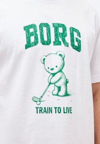 Λευκό πουκάμισο με πράσινο κείμενο "BORG" και "TRAIN TO LIVE" που δείχνει μια αρκουδάκι να κρατά ένα γκολφ ρόπαλο, έτοιμο να χτυπήσει μια μπάλα του γκολφ.