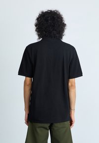 Polo noir à manches courtes, texture en coton lisse, à col. Associé à un pantalon vert olive, vu de dos.