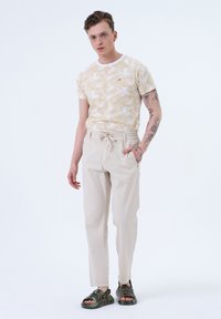 T-shirt à manches courtes avec un motif de feuilles de palmier jaune et blanc, associé à un pantalon beige fuselé et des sandales vert foncé.