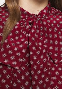 Primer plano de una persona que lleva una blusa burdeos con un patrón geométrico y un gran lazo atado en el cuello.