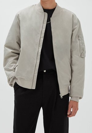Personne portant une veste bomber gris clair avec une poche zippée sur la manche gauche, une chemise noire et un pantalon noir sur un fond uni.