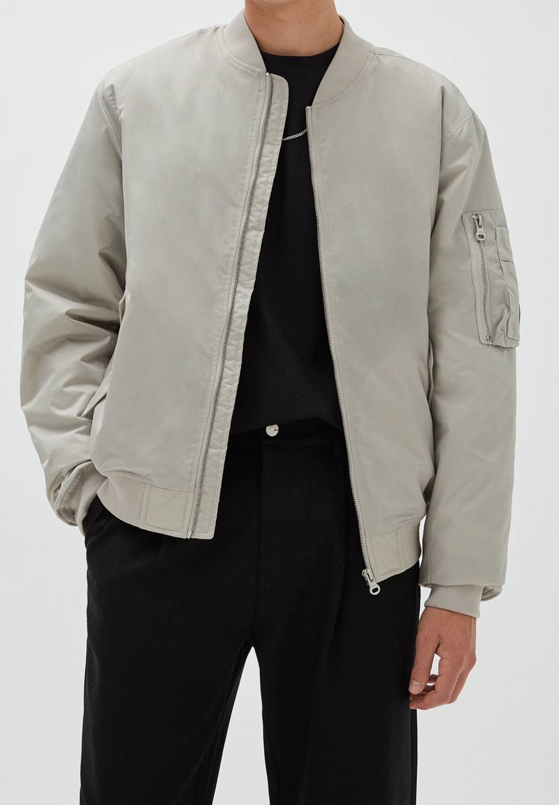 Personne portant une veste bomber gris clair avec une poche zippée sur la manche gauche, une chemise noire et un pantalon noir sur un fond uni.
