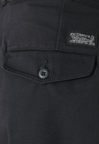 Tela de denim negro con un cierre de botón visible y un bolsillo con solapa. Una etiqueta de Levi Strauss & Co. está cosida en el bolsillo.