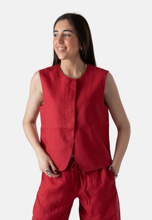 Mujer sonriendo y mirando hacia la izquierda, vestida con una blusa roja sin mangas y pantalones rojos a juego con cordón, manos en los bolsillos, sobre un fondo liso.