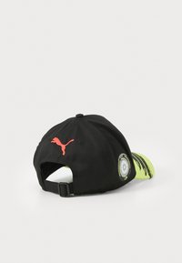 Svart caps med justerbar stropp, med en rød logo og en neongul underside, som viser et grafisk trykk på skyggen.