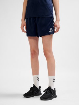 Person iført marineblå sportsshorts og top, hvide sokker med sorte chevroner og sorte atletiksk sko, står på en ensfarvet baggrund.