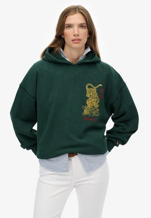 Donkergroene oversized hoodie van zacht materiaal, met een gouden draakafbeelding en geborduurde tekst op de voorkant. Gelaagd over een blauwgestreept shirt.