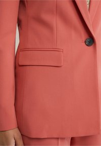 Detailní záběr osoby v korálovém saku s černým knoflíkem a kapsou na klopě na levé straně, paže částečně viditelná.