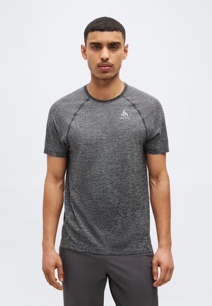 Homme debout de face portant un t-shirt de sport texturé gris à manches courtes et un pantalon gris foncé devant un fond blanc uni.