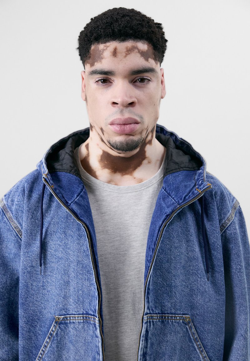 Giubbotto di jeans blu con cappuccio, chiusura con zip e tasche frontali, indossato sopra una maglietta grigia chiara. Il modello presenta un motivo della pelle unico.