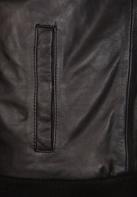 Zwart leren materiaal met een gladde textuur, met een rechthoekige zak. De onderkant heeft een ribgebreide accent, wat contrast toevoegt.