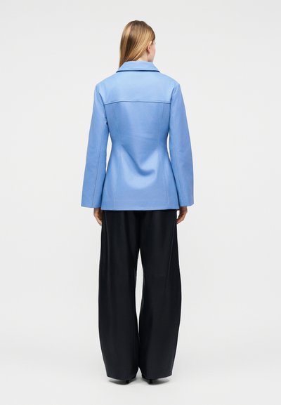 Γυναίκα με μακριά ξανθά μαλλιά, που φοράει εφαρμοστή ανοιχτόγαλανη ζακέτα και φαρδιά μαύρη παντελόνα, όρθια με την πλάτη προς τα εμπρός σε λευκό φόντο.