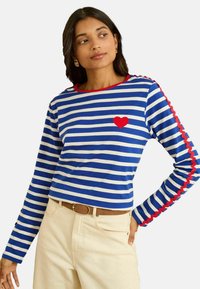 Blau-weiß gestreiftes Langarmshirt mit rotem, gezacktem Saum und einem roten Herzaufnäher auf der linken Brust. Kombiniert mit hellfarbiger Hose.