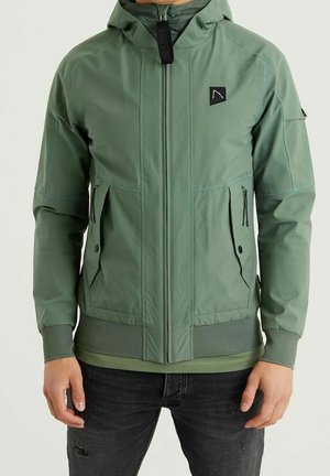 Homme portant une veste à capuche vert clair avec fermeture éclair et poches à boutons, poignets et ourlet côtelés, associée à un jean noir déchiré.