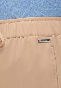 Ανοιχτό μπεζ σορτς με ελαστική μέση, κορδόνι για κλείσιμο και ασημένια ετικέτα με το λογότυπο "Calvin Klein". Λείο ύφασμα, μινιμαλιστικός σχεδιασμός.