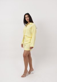 Mujer que lleva una camisa amarilla de manga larga con botones y shorts a juego con sandalias transparentes de tacón alto, de pie frente a un fondo blanco.