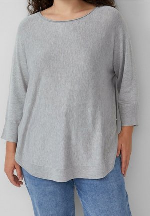 Femme portant un pull en maille gris clair ample à manches trois-quarts et un jean bleu, debout devant un fond uni.