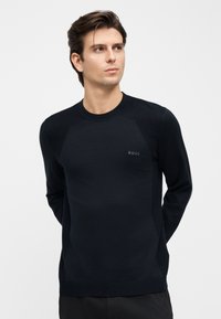 Mladý muž v přiléhavém černém svetru BOSS, stojící s rukama za zády, mírně hledící doprava na jednoduchém světlém pozadí.