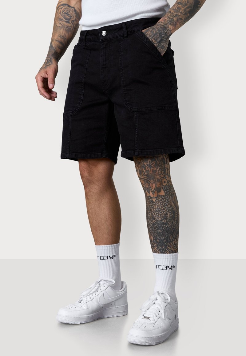 A man stands wearing black cargo shorts, white socks, and white sneakers, showcasing intricate tattoos on his right leg.