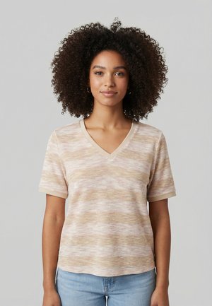 Femme aux cheveux bouclés portant un haut à manches courtes à rayures beige et blanches avec col en V, et un jean bleu clair, debout devant un fond gris uni.
