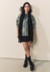 Grå og grøn argyle sweater med knapper, sort tørklæde med frynser, sort nederdel og knæhøje sorte støvler med spænder.