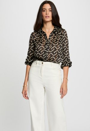 Blouse noire transparente avec un motif ondulé blanc et beige, dotée d'un devant boutonnière et de manches retroussées, assortie à un pantalon large blanc.