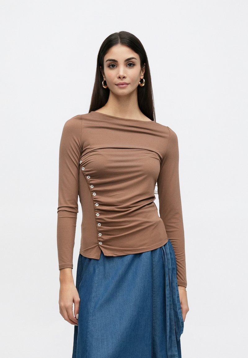 Pruun pika varrukaga pluus laia kaelusega, kokku kistud kangast detailide ja dekoratiivsete nuppudega külje peal. Kandmisel teksaseelikuga.